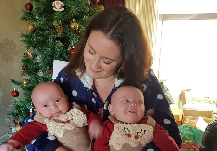 A woman holds two babies in her arms, all facing the camera, looking down at the baby on the left