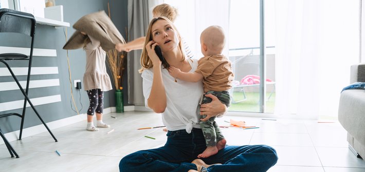 A woman sits cross legged on a floor with a phone to her ear in one hand and a baby in her other, whilst two children fight with cushions in the background
