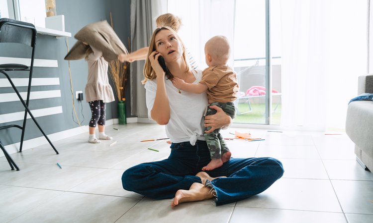 A woman sits cross legged on a floor with a phone to her ear in one hand and a baby in her other, whilst two children fight with cushions in the background