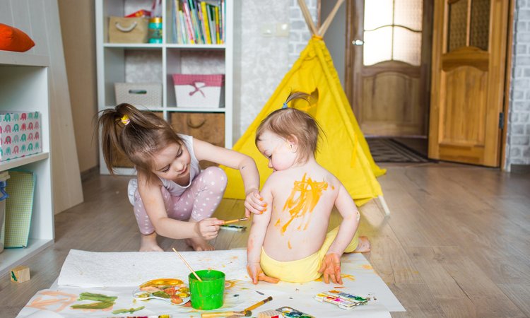 Two young girls sit on the floor as one paints the back of the other