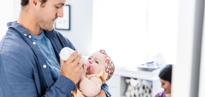 A man in the foreground bottle feeds a baby as a woman in the background does the same