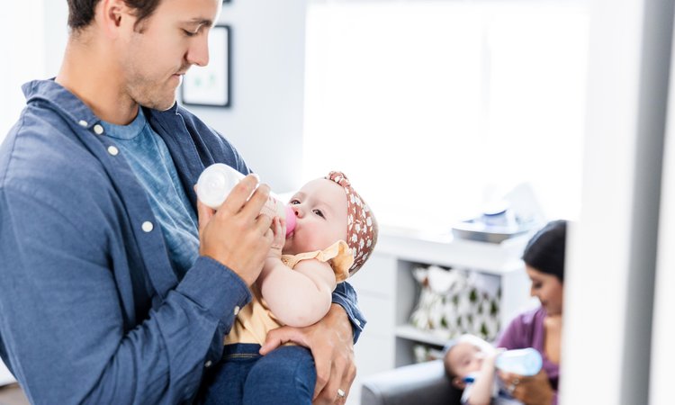 A man in the foreground bottle feeds a baby as a woman in the background does the same