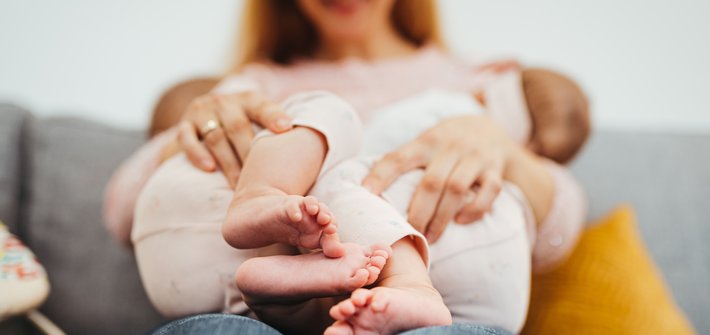 A woman in the background holds two babies in the foreground who are breastfeeding