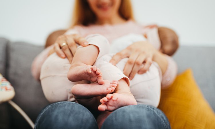 A woman in the background holds two babies in the foreground who are breastfeeding