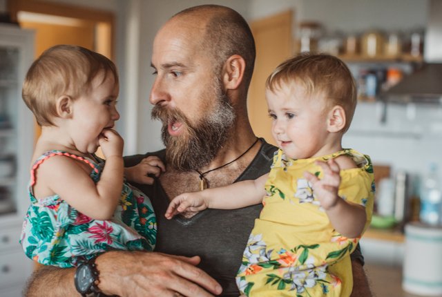 A man holds two toddlers in his arms as he makes a face at one of them