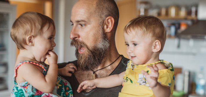 A man holds two toddlers in his arms as he makes a face at one of them