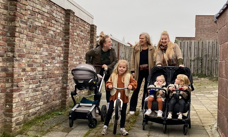 Three mothers walk along a path, two pushing buggies whilst a girl rides a bike in between them