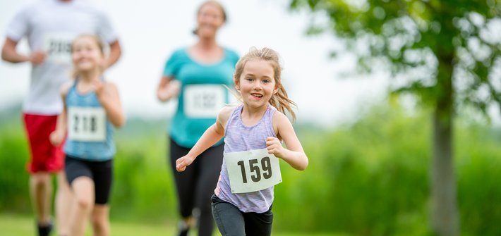 A young girl in the foreground runs towards the camera as a man, woman and child in the background chase after her