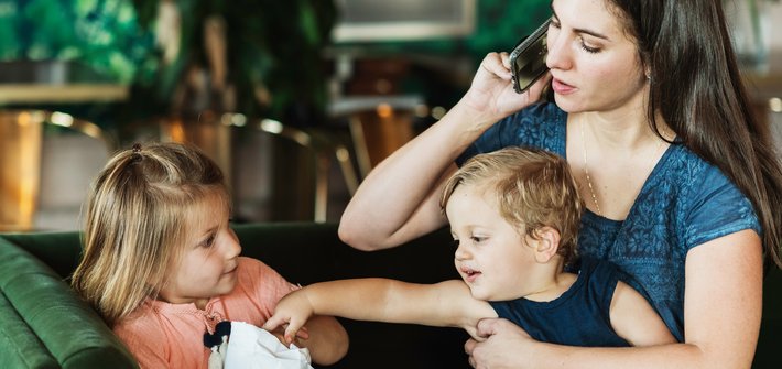 A woman talks on the phone in one hand whilst holding a boy on her lap in the other who is reaching out trying to take something from a girl sat close by