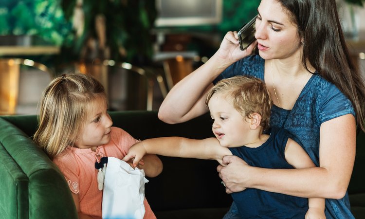 A woman talks on the phone in one hand whilst holding a boy on her lap in the other who is reaching out trying to take something from a girl sat close by