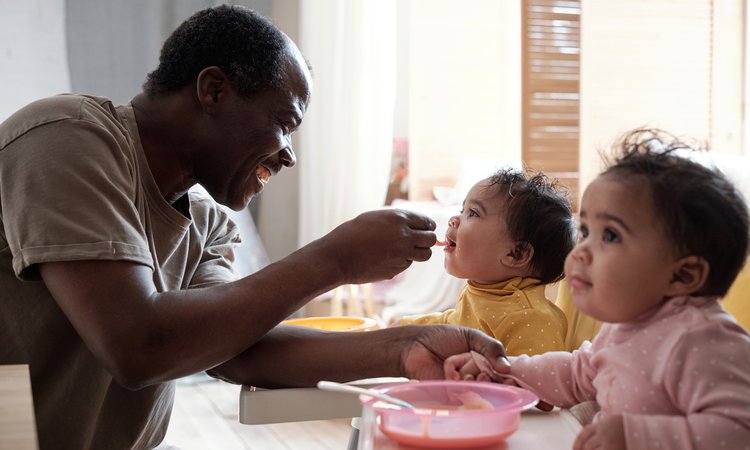 Two toddlers are sat at a table with a bowl in front of them as a man spoon feeds one of the toddlers