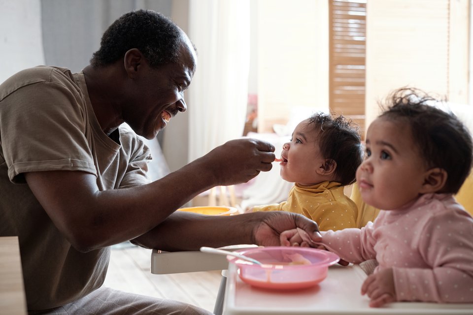 Two toddlers are sat at a table with a bowl in front of them as a man spoon feeds one of the toddlers