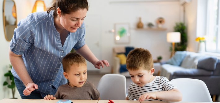 A set of twins sit playing at a table as a woman leans over to offer support