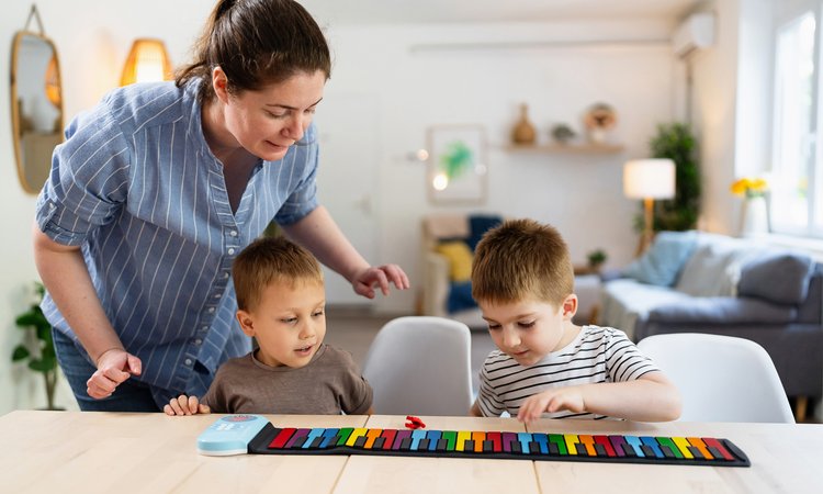 A set of twins sit playing at a table as a woman leans over to offer support