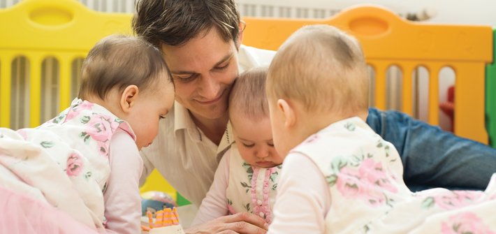 A man lays on a bed taking care of three triplets who sit playing in front of him