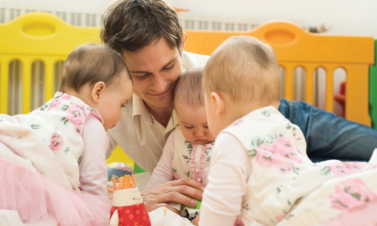 A man lays on a bed taking care of three triplets who sit playing in front of him