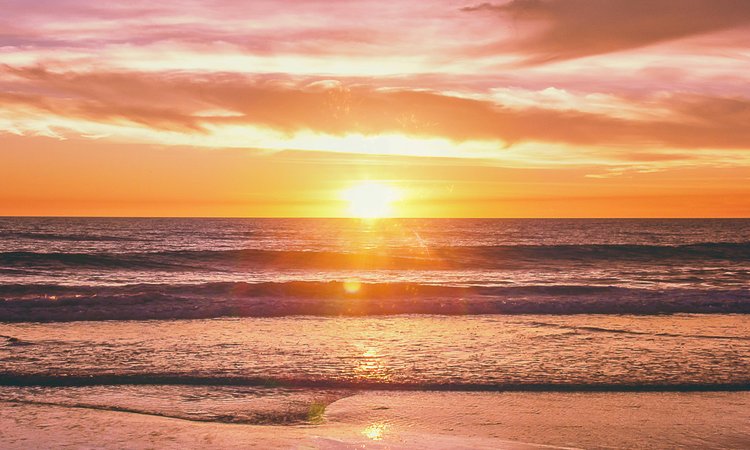 A sunset in the distance with waves rolling onto a beach in the foreground
