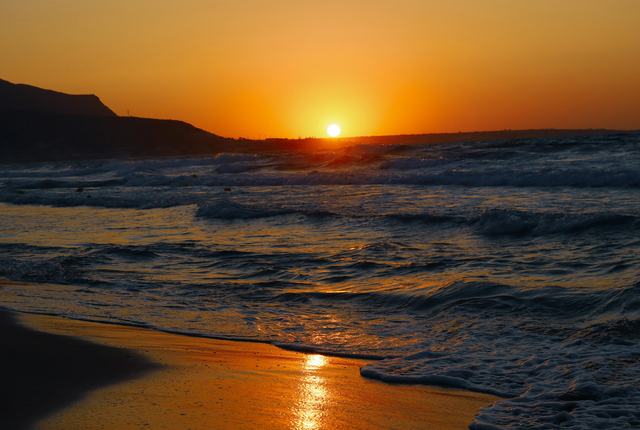 A sunset in the distance with waves rolling onto a beach in the foreground