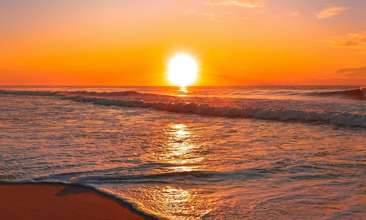 A sunset in the distance with waves rolling onto a beach in the foreground