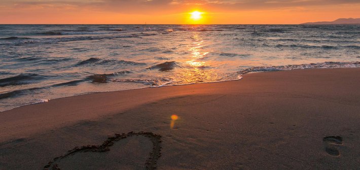 A sunset over the ocean in the distance with a beach in the foreground and a heart shape drawn into the sand