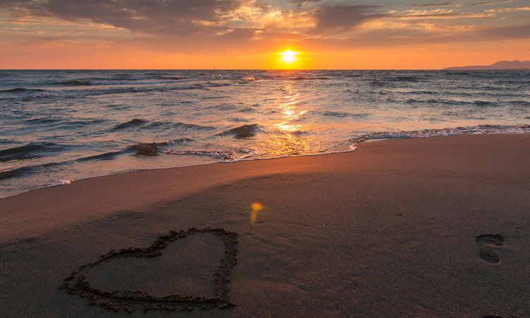 A sunset over the ocean in the distance with a beach in the foreground and a heart shape drawn into the sand