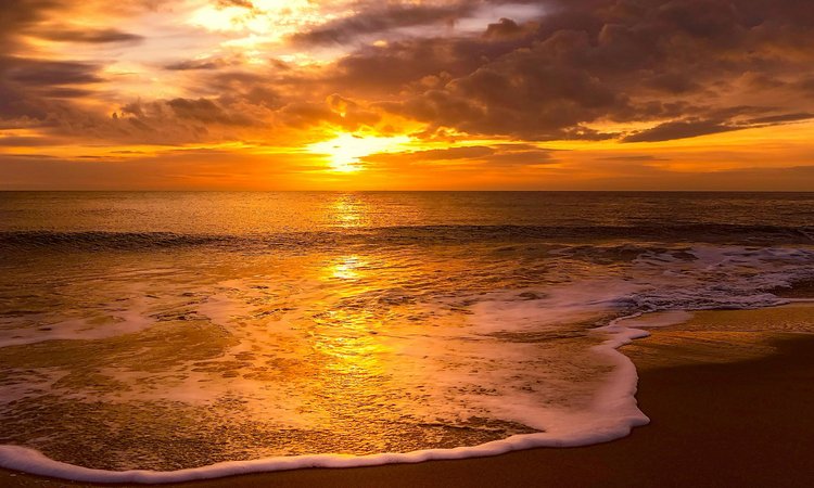 A sunset in the distance with waves rolling onto a beach in the foreground