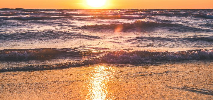 A sunset in the distance with waves rolling onto a beach in the foreground