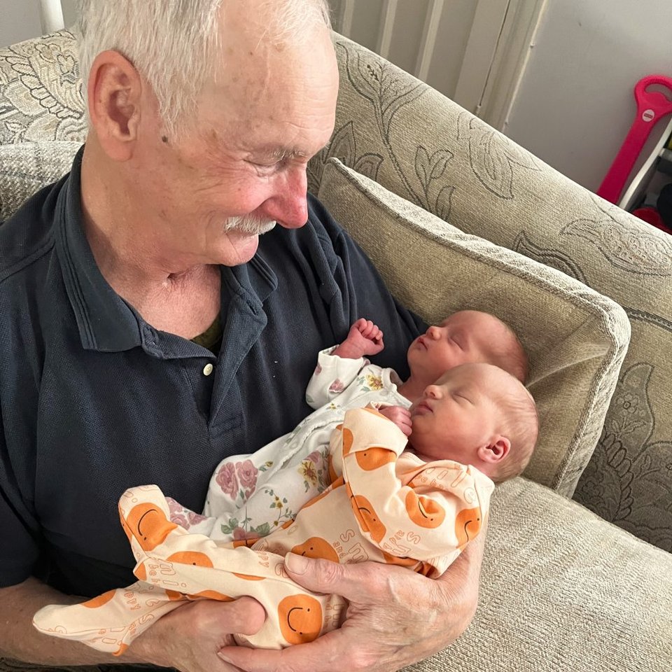 an elderly man sat on a sofa holding a set of twin babies