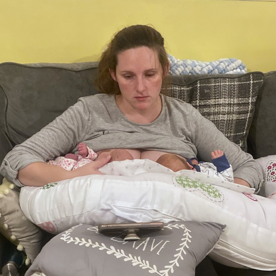 woman tandem breastfeeding her twins resting on cushions while watching media on her smartphone
