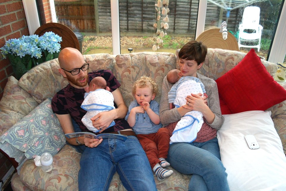 twin babies held by their mother and father who is reading a story with an older older child sitting in the middle