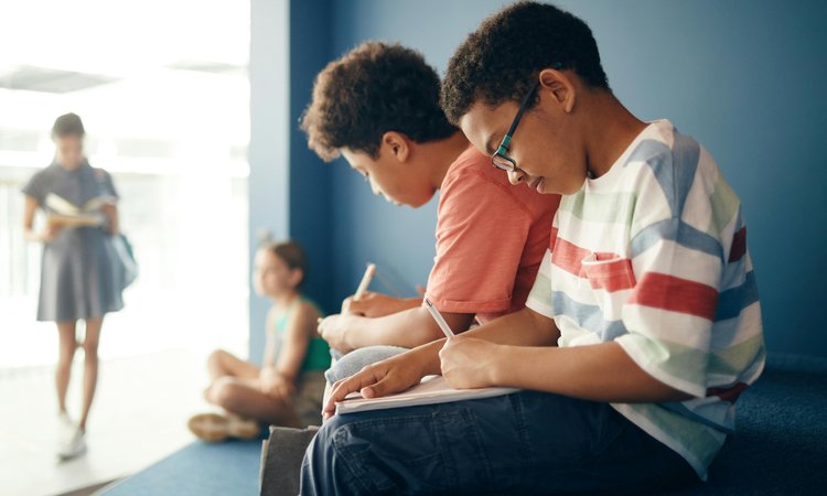Two boys sit next to each other writing separately in notebooks