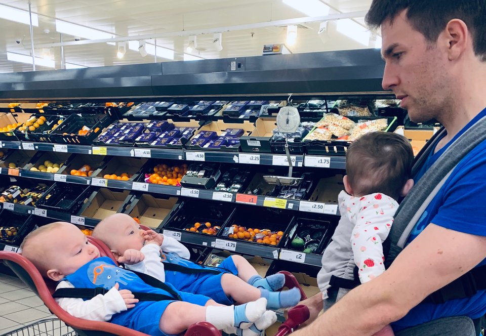 A man stands in a supermarket grocery aisle with two babies in a twin baby trolly and wearing a third baby