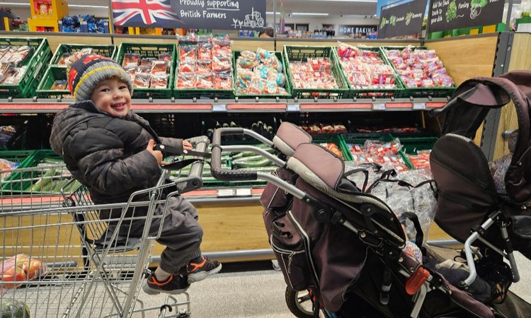 A baby sits in a twin trolly on the left with a twin buggy to the right
