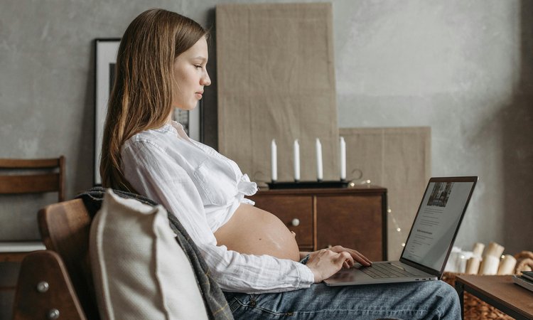 A pregnant woman sits on a sofa typing on a laptop, viewed from the side
