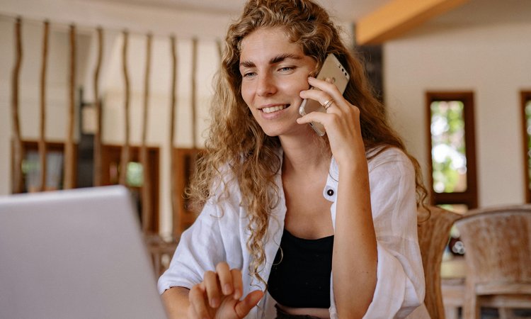 A woman holds a phone to her ear whilst smiling and looking at a laptop