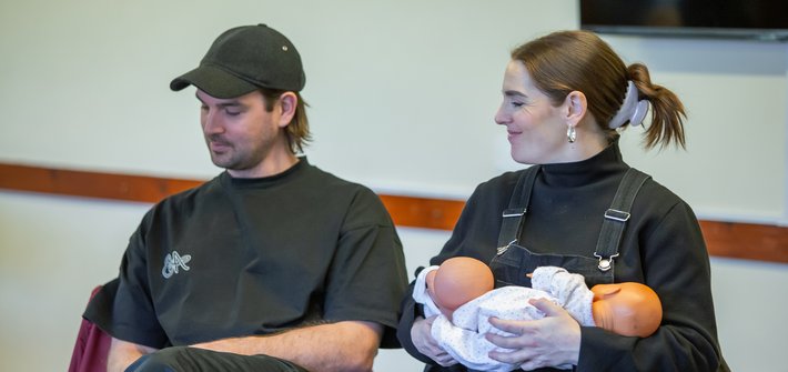A man and a woman sit together with the woman holding two toy babies in her arms