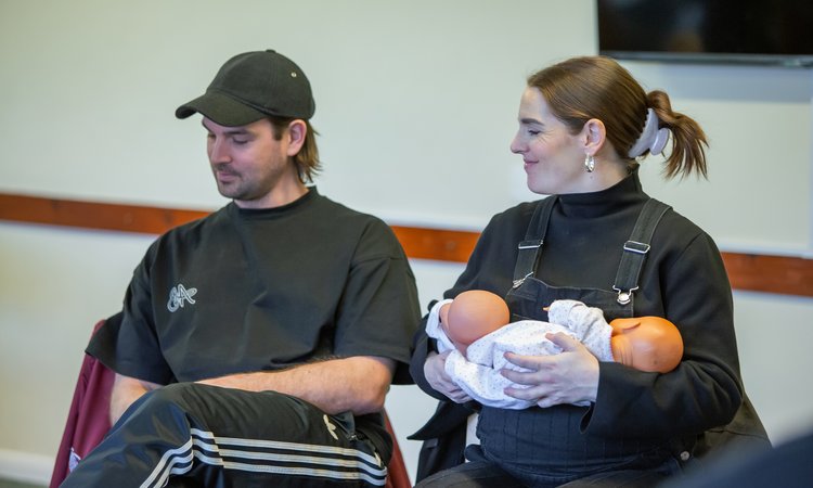 A man and a woman sit together with the woman holding two toy babies in her arms