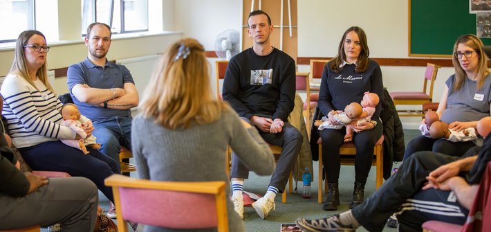 A group of people sit in a circle looking at a teacher in the foreground with their back to the camera
