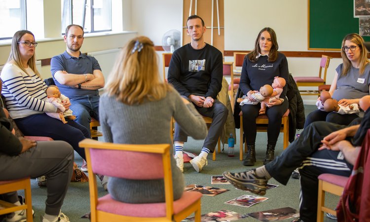 A group of people sit in a circle looking at a teacher in the foreground with their back to the camera