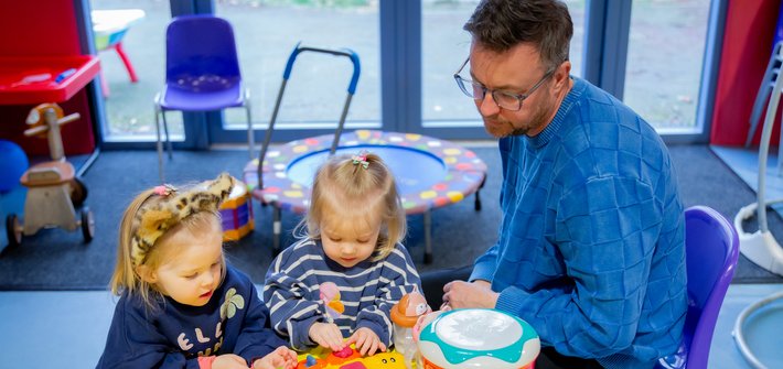 A man sits at a table with young twin girls playing with toys