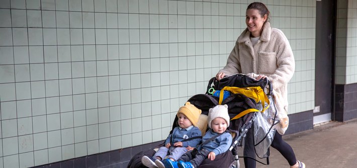 A woman pushes a twin pushchair with twin boys sat in it