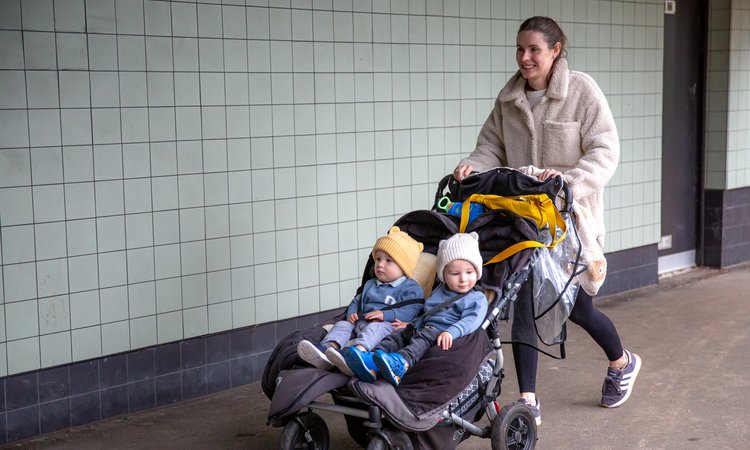 A woman pushes a twin pushchair with twin boys sat in it