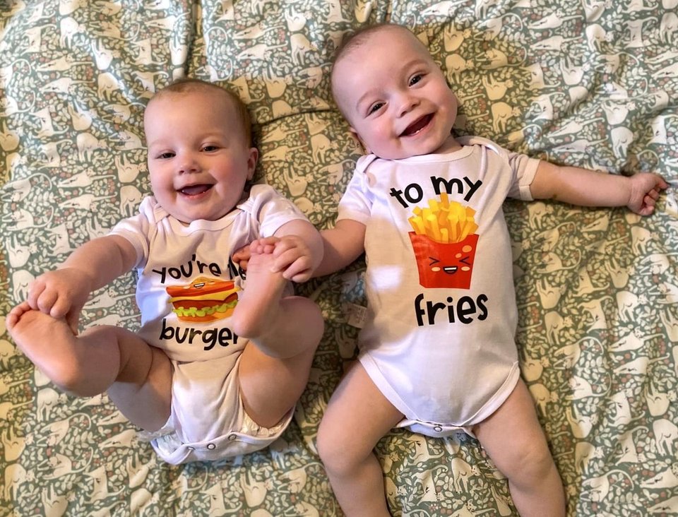 twin babies smiling lying on a bed, one wearing a onesie reading 'you're the burger' and the other baby wearing a onesie reading 'to my fries'