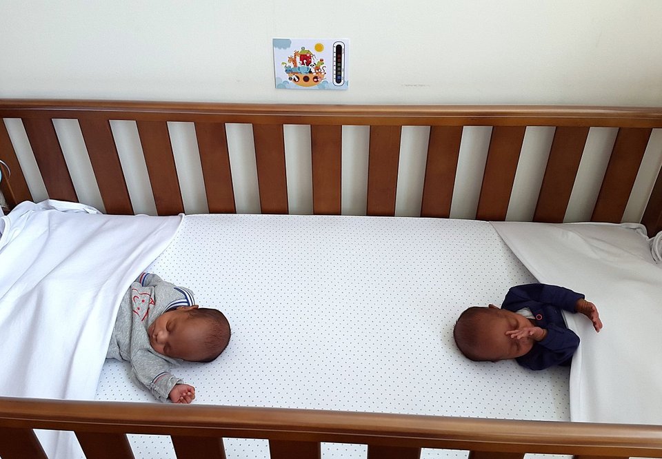 twin babies sleeping on either end of a baby cot with a thermometer on stuck on the wall next to the cot