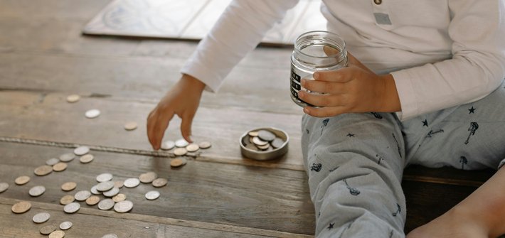 A child sits on the floor picking up coins spread across the floor and putting them into a glass jar in their other hand