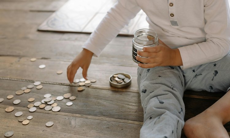 A child sits on the floor picking up coins spread across the floor and putting them into a glass jar in their other hand