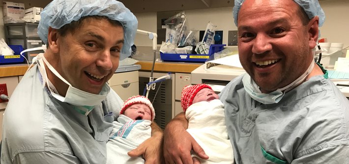 Two men dressed in hospital scrubs smile at the camera as they each cradle a baby