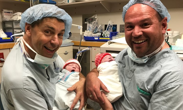 Two men dressed in hospital scrubs smile at the camera as they each cradle a baby