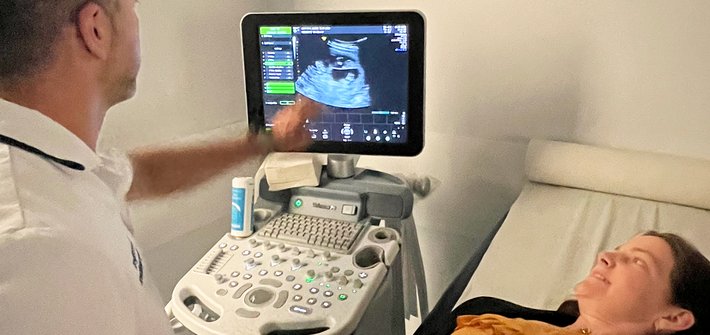 Looking over the shoulder of a sonographer pointing at an ultrasound machine with a woman lying next to him