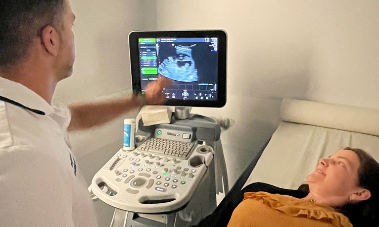 Looking over the shoulder of a sonographer pointing at an ultrasound machine with a woman lying next to him